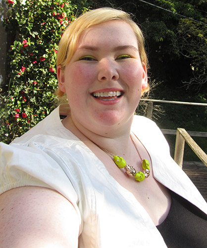 Me (a fat, blonde white woman) doing a selfie with a big smile on my face.  I'm wearing neon yellow eyeshadow and a necklace of neon yellow and gold skulls.  A little of my white short-sleeved jacket and black scoop neck dress can also be seen.  There is a camellia bush in the background.